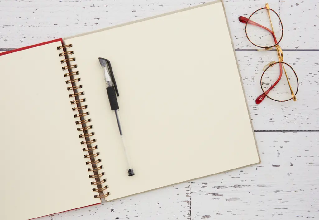 A notebook lying open beside a pen and reading glasses on a pale table