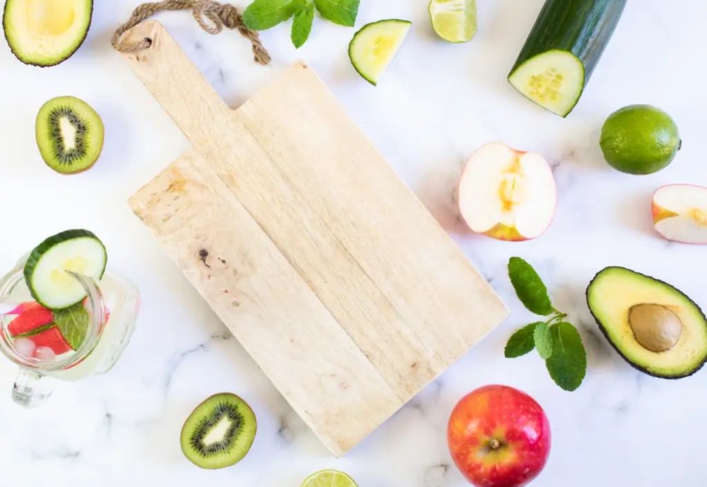 A wooden chopping board surrounded by fresh vegetables and fruit
